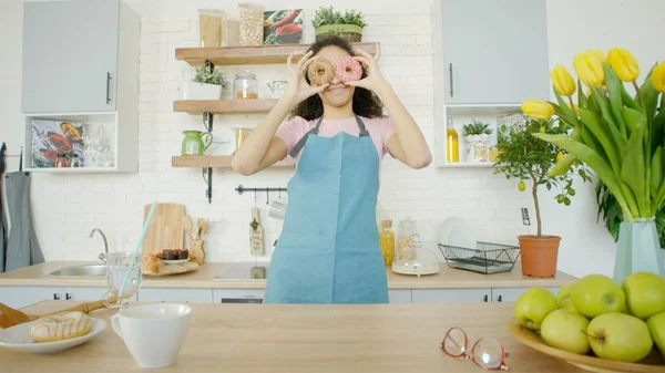 A fiatal nő fánkkal a kezében mászkál a konyhában. Stock Kép