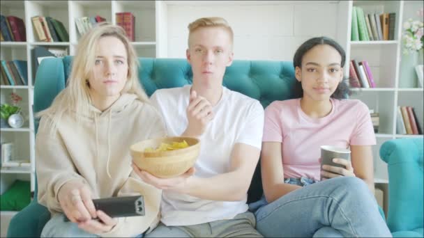 Feliz tres amigos están viendo la televisión y comiendo bocadillos juntos en el sofá — Vídeos de Stock