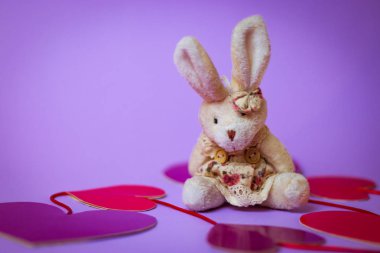 Oyuncak tavşan kağıt kalpler mor bir arka plan üzerinde kırmızı kurdele ile beyaz hediye kutusu üzerinde oturur.