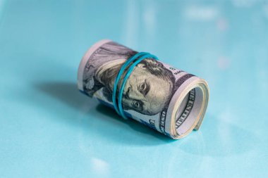 Yuvarlanmış bir tomar paranın yakın plan fotoğrafı. Kenarında duran mavi lastik bantla Benjamin Franklin 'in yüzüne dolanan 100 dolarlık banknotla fotokopi çekilen beyaz arka planda..