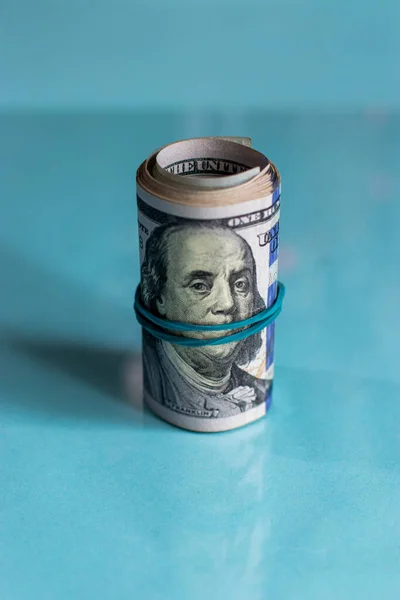 Yuvarlanmış bir tomar paranın yakın plan fotoğrafı. Kenarında duran mavi lastik bantla Benjamin Franklin 'in yüzüne dolanan 100 dolarlık banknotla fotokopi çekilen beyaz arka planda..