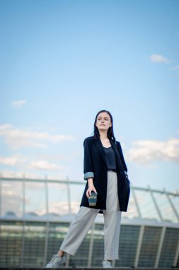 Genç güzel kız elinde bir fincan kahveyle sokakta yürüyor, güneşli bir günde.