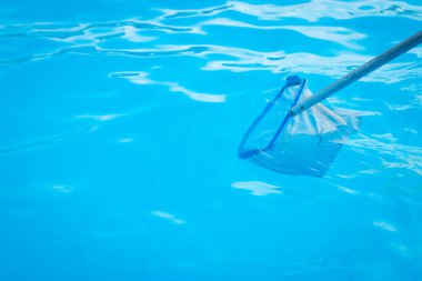 Arka bahçeye yerleştirilmiş portatif bir yüzme havuzunun dibini teleskopik bir direğe monte edilmiş fırçayla temizleyen genç bir beyaz adamın yakın çekimi.