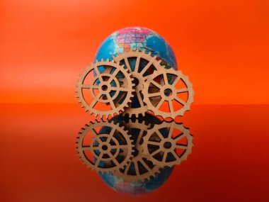 Küre ahşap dişlilerle çevrili, turuncu arka planda yansıtıcı bir yüzey üzerinde dinleniyor..