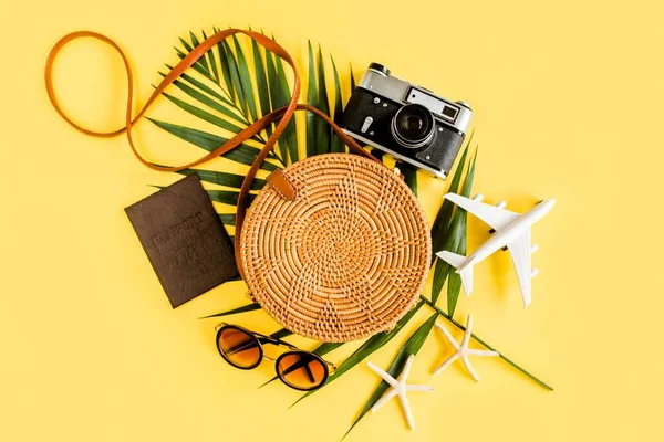 Traveler accessori concetto su sfondo giallo. Borsa rotonda in rattan naturale fatto a mano alla moda, fotocamera retrò e foglie tropicali. — Foto Stock