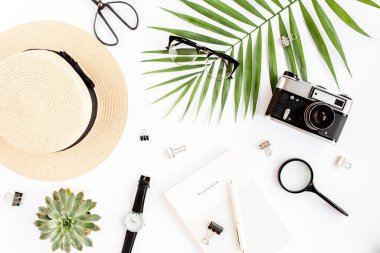 Üst kat ofis masası. Hasır şapka, gözlük, retro kamera, not defteri ve beyaz arka planda tropikal palmiye yaprağı olan çalışma alanı. Düz yatış, üst görünüm.