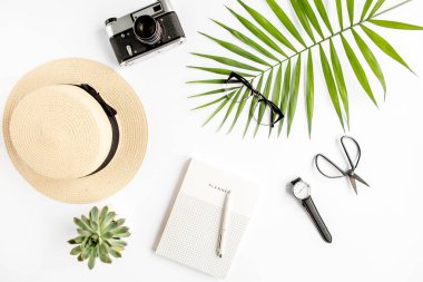 Üst kat ofis masası. Hasır şapka, gözlük, retro kamera, not defteri ve beyaz arka planda tropikal palmiye yaprağı olan çalışma alanı. Düz yatış, üst görünüm.