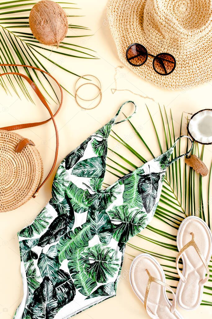 Womans beach accessories: swimsuit with tropical print, rattan bag, straw hat, tropical palm leaves on yellow background. Summer background. 