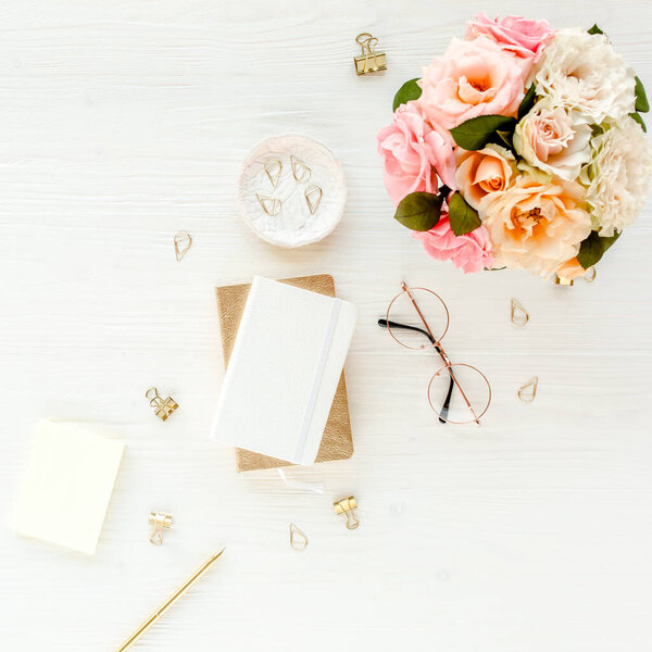 Flat lay womens office desk. Female workspace with pink roses flowers bouquet, accessories, golden diary, glasses on white wooden background Top view