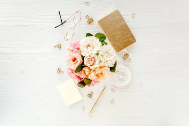 Düz uzanmış kadınların ofis masası. Pembe güller, çiçek buketi, aksesuarlar, altın günlük, beyaz ahşap arka planda gözlük.