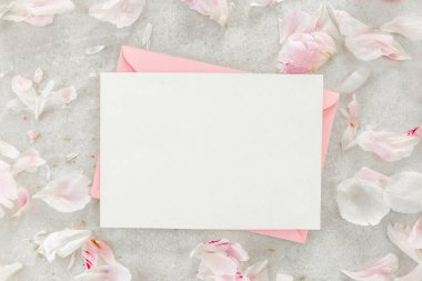 Sahte davetiye, boş kağıt tebrik kartı, pembe zarf ve gri taş masada şakayıklar. Çiçek arkaplanı. Düz yatış, üst görünüm.