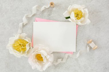 Sahte davetiye, boş kağıt tebrik kartı, pembe zarf ve gri taş masada şakayıklar. Çiçek arkaplanı. Düz yatış, üst görünüm.