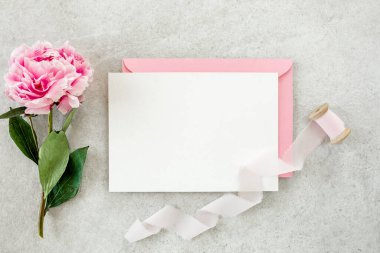 Sahte davetiye, boş kağıt tebrik kartı, pembe zarf ve gri taş masada şakayıklar. Çiçek arkaplanı. Düz yatış, üst görünüm.
