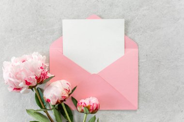 Sahte davetiye, boş kağıt tebrik kartı, pembe zarf ve gri taş masada şakayıklar. Çiçek arkaplanı. Düz yatış, üst görünüm.
