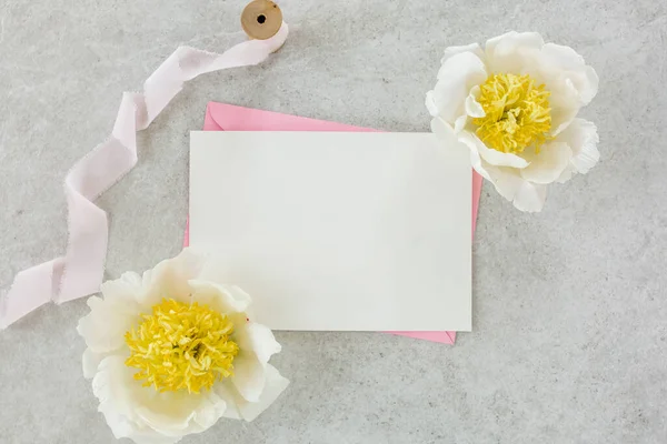 Mockup invitation, blank paper greeting card, pink envelope and peonies on gray stone table. Flower background. Flat lay, top view. — Stock Photo, Image