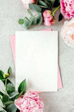 Sahte davetiye, boş kağıt tebrik kartı, pembe zarf ve gri taş masada şakayıklar. Çiçek arkaplanı. Düz yatış, üst görünüm.