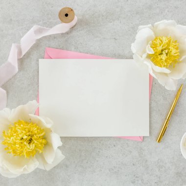 Sahte davetiye, boş kağıt tebrik kartı, pembe zarf ve gri taş masada şakayıklar. Çiçek arkaplanı. Düz yatış, üst görünüm.