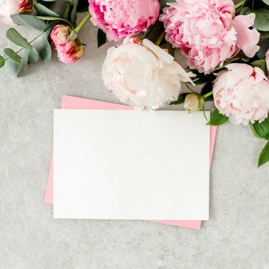 Sahte davetiye, boş kağıt tebrik kartı, pembe zarf ve gri taş masada şakayıklar. Çiçek arkaplanı. Düz yatış, üst görünüm.
