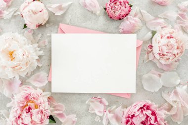 Sahte davetiye, boş kağıt tebrik kartı, pembe zarf ve gri taş masada şakayıklar. Çiçek arkaplanı. Düz yatış, üst görünüm.