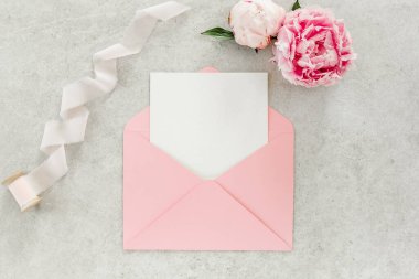 Sahte davetiye, boş kağıt tebrik kartı, pembe zarf ve gri taş masada şakayıklar. Çiçek arkaplanı. Düz yatış, üst görünüm.