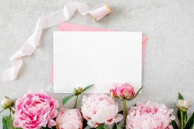 Sahte davetiye, boş kağıt tebrik kartı, pembe zarf ve gri taş masada şakayıklar. Çiçek arkaplanı. Düz yatış, üst görünüm.