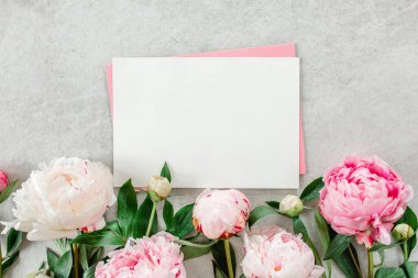 Sahte davetiye, boş kağıt tebrik kartı, pembe zarf ve gri taş masada şakayıklar. Çiçek arkaplanı. Düz yatış, üst görünüm.