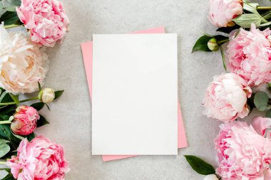 Sahte davetiye, boş kağıt tebrik kartı, pembe zarf ve gri taş masada şakayıklar. Çiçek arkaplanı. Düz yatış, üst görünüm.