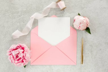 Sahte davetiye, boş kağıt tebrik kartı, pembe zarf ve gri taş masada şakayıklar. Çiçek arkaplanı. Düz yatış, üst görünüm.