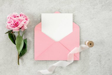 Sahte davetiye, boş kağıt tebrik kartı, pembe zarf ve gri taş masada şakayıklar. Çiçek arkaplanı. Düz yatış, üst görünüm.