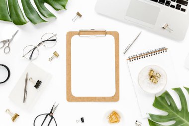 Ev ofisinin çalışma alanı dizüstü bilgisayarı, tropikal yaprakları Monstera, not panosu, defter ve aksesuarları beyaz arka planda. Düz yatış, üst görünüm