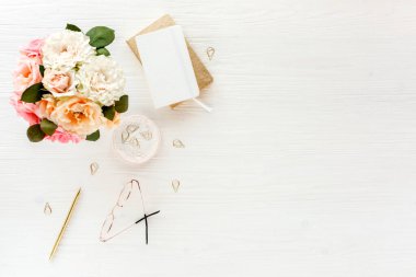 Düz uzanmış kadınların ofis masası. Pembe güller, çiçek buketi, aksesuarlar, altın günlük, beyaz ahşap arka planda gözlük.