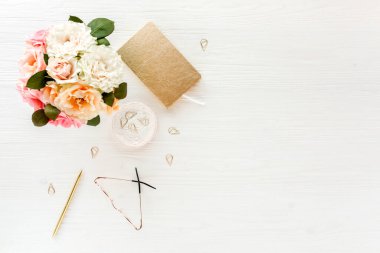 Düz uzanmış kadınların ofis masası. Pembe güller, çiçek buketi, aksesuarlar, altın günlük, beyaz ahşap arka planda gözlük.