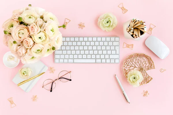 Tarz sahibi, pembe kadınların ofis masası. Bilgisayarlı çalışma alanı, buket ranunculus ve güller, pembe arka planda pano. Düz yatıyordu. Üst görünüm.