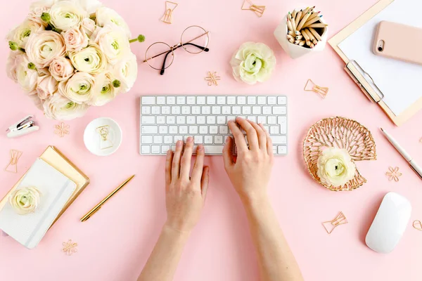Stilisierter, pinkfarbener Home-Office-Schreibtisch für Frauen. Arbeitsplatz mit weiblichen Händen, Computer, Strauß Hahnenfuß und Rosen, Klemmbrett auf rosa Hintergrund. Flache Lage. — Stockfoto