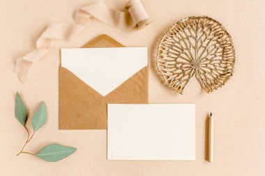 Sahte düğün davetiyesi, boş tebrik kartı ve el işi zarfı, yeşil yapraklı okaliptüs. Düz yatış, üst görünüm.