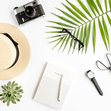 Üst kat ofis masası. Hasır şapka, gözlük, retro kamera, not defteri ve beyaz arka planda tropikal palmiye yaprağı olan çalışma alanı. Düz yatış, üst görünüm.