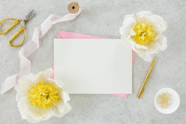 Sahte davetiye, boş kağıt tebrik kartı, pembe zarf ve gri taş masada şakayıklar. Çiçek arkaplanı. Düz yatış, üst görünüm.