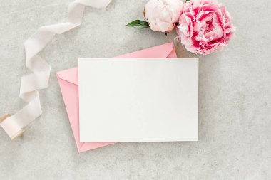 Sahte davetiye, boş kağıt tebrik kartı, pembe zarf ve gri taş masada şakayıklar. Çiçek arkaplanı. Düz yatış, üst görünüm.