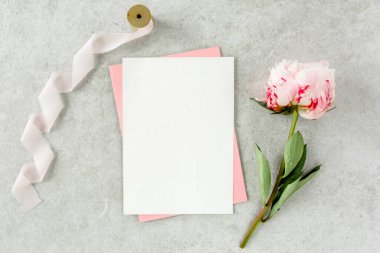Sahte davetiye, boş kağıt tebrik kartı, pembe zarf ve gri taş masada şakayıklar. Çiçek arkaplanı. Düz yatış, üst görünüm.