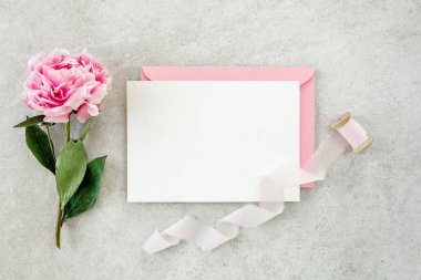 Sahte davetiye, boş kağıt tebrik kartı, pembe zarf ve gri taş masada şakayıklar. Çiçek arkaplanı. Düz yatış, üst görünüm.