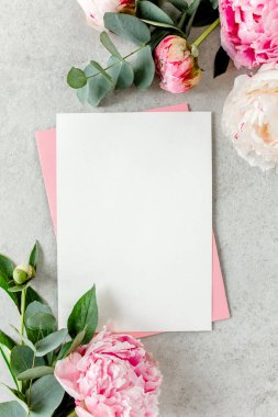 Sahte davetiye, boş kağıt tebrik kartı, pembe zarf ve gri taş masada şakayıklar. Çiçek arkaplanı. Düz yatış, üst görünüm.