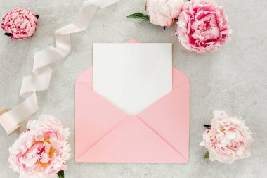 Sahte davetiye, boş kağıt tebrik kartı, pembe zarf ve gri taş masada şakayıklar. Çiçek arkaplanı. Düz yatış, üst görünüm.