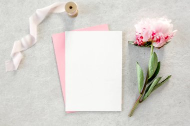 Sahte davetiye, boş kağıt tebrik kartı, pembe zarf ve gri taş masada şakayıklar. Çiçek arkaplanı. Düz yatış, üst görünüm.
