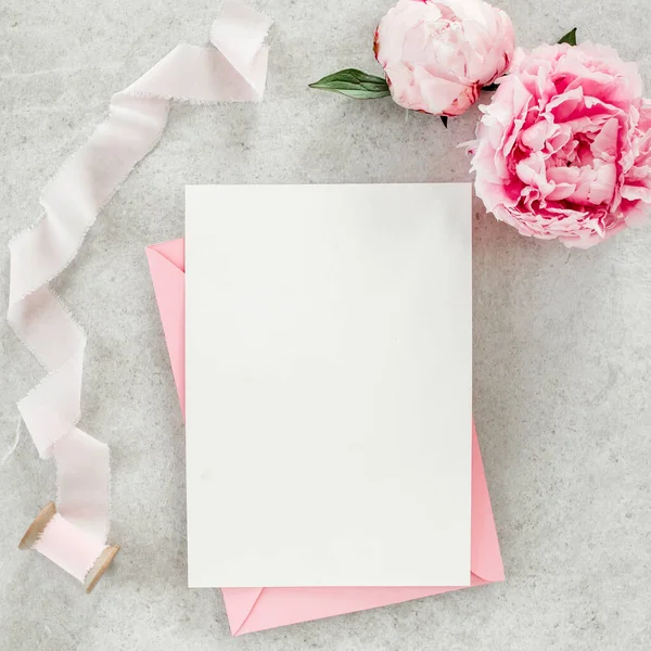 Invitation maquette, carte de vœux en papier vierge, enveloppe rose et pivoines sur table en pierre grise. Fond de fleur. Couché plat, vue du dessus. — Photo