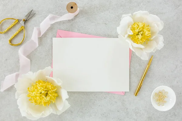 Sahte davetiye, boş kağıt tebrik kartı, pembe zarf ve gri taş masada şakayıklar. Çiçek arkaplanı. Düz yatış, üst görünüm.