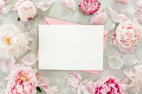 Sahte davetiye, boş kağıt tebrik kartı, pembe zarf ve gri taş masada şakayıklar. Çiçek arkaplanı. Düz yatış, üst görünüm.
