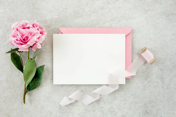 Sahte davetiye, boş kağıt tebrik kartı, pembe zarf ve gri taş masada şakayıklar. Çiçek arkaplanı. Düz yatış, üst görünüm.