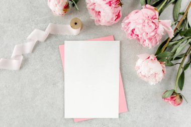 Sahte davetiye, boş kağıt tebrik kartı, pembe zarf ve gri taş masada şakayıklar. Çiçek arkaplanı. Düz yatış, üst görünüm.