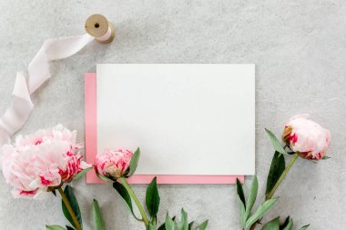 Sahte davetiye, boş kağıt tebrik kartı, pembe zarf ve gri taş masada şakayıklar. Çiçek arkaplanı. Düz yatış, üst görünüm.