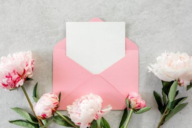 Sahte davetiye, boş kağıt tebrik kartı, pembe zarf ve gri taş masada şakayıklar. Çiçek arkaplanı. Düz yatış, üst görünüm.